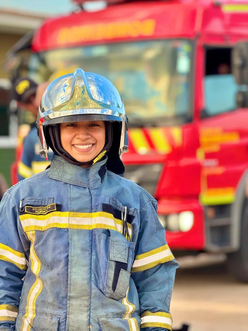 Bombeira sorrindo, de capacete e farda, diante de um caminhão de bombeiros